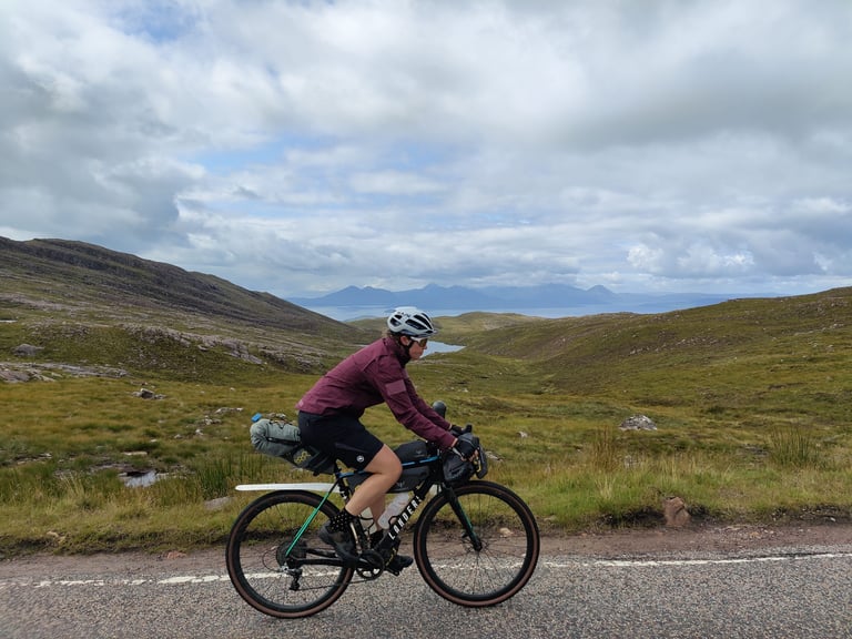 Laura Fidler rides a loaded gravel bike along a road in the Scottish Highlands, Apidura Downtube Pack on board.