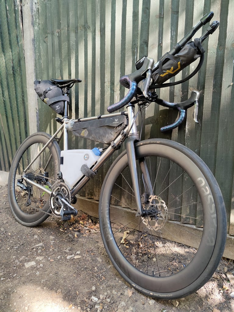 Chris Herbert's drop-bar bike with an Apidura Downtube Pack strapped between the aerobars, against a green fence.