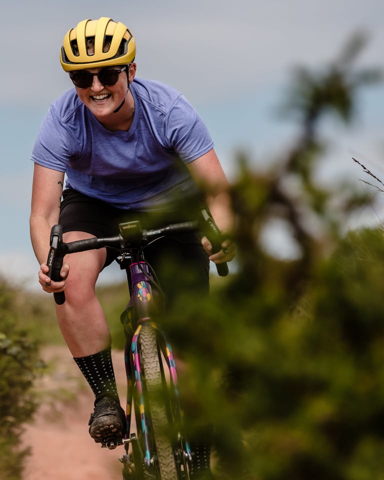 Rider in a yellow helmet grins as she rides a colourful gravel bike on WTB Nano SG2 tyres past gorse.