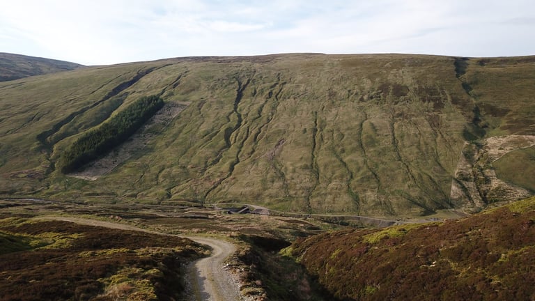 Aerial view of a gravel track winding through heather below a steep grassy hillside in Highland Perthshire.