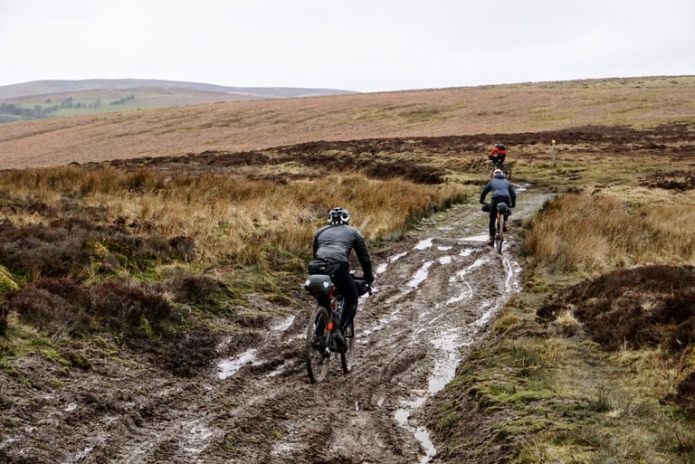 Winter bikepackers ride loaded bikes along a rutted, flooded track across open moorland.