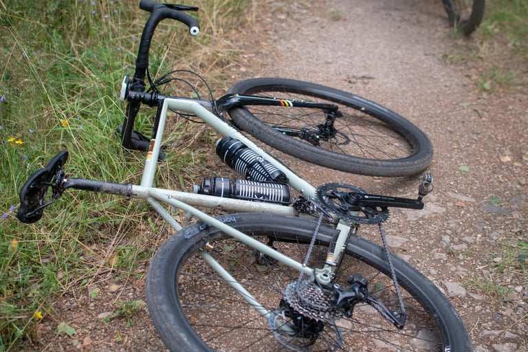 Sage green Bombtrack Audax lying on its side on a dry dirt path beside long grass, two bottles in its cages.