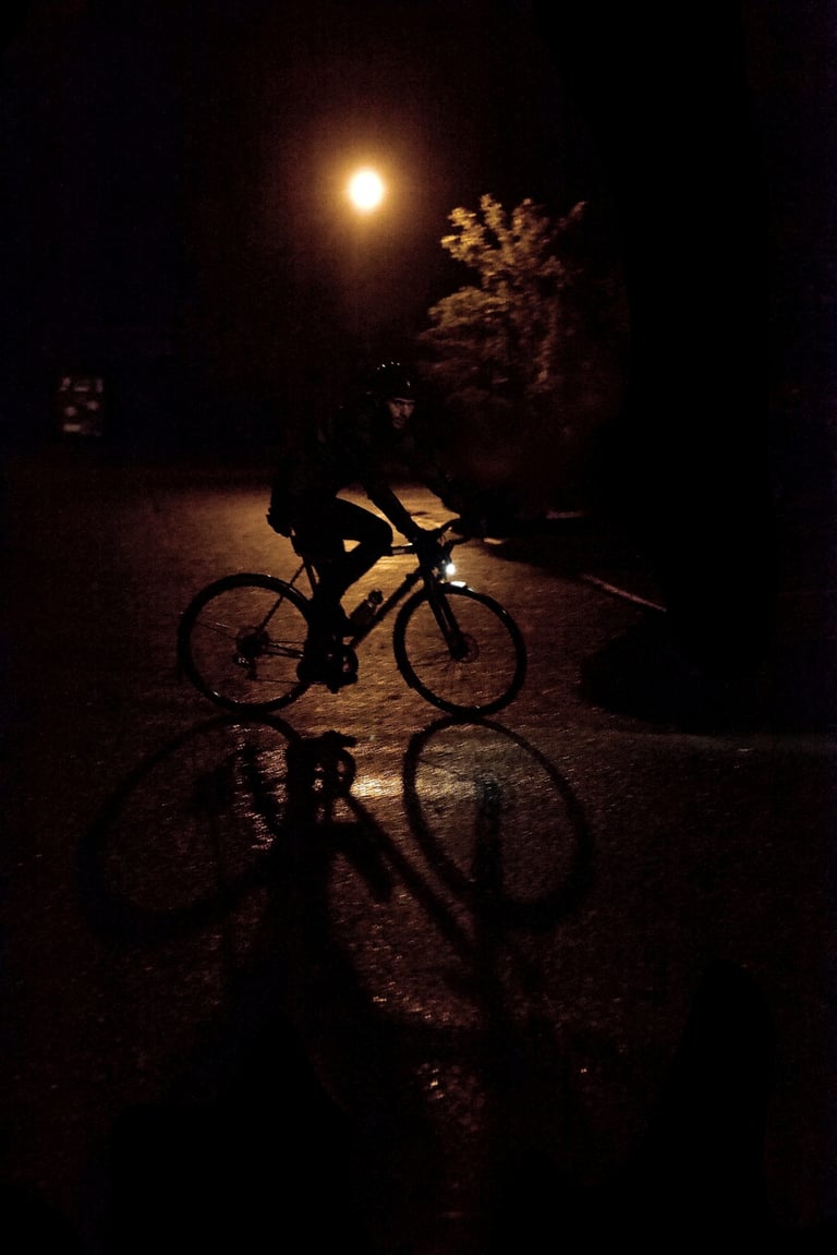 Cyclist rides a dynamo-lit bike through a dark, wet street at night, a street lamp glowing behind.
