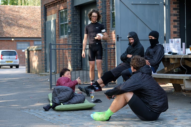 Riders sit and talk outside a brick building at the Great British Escapade finish, one lying on a sleeping mat.