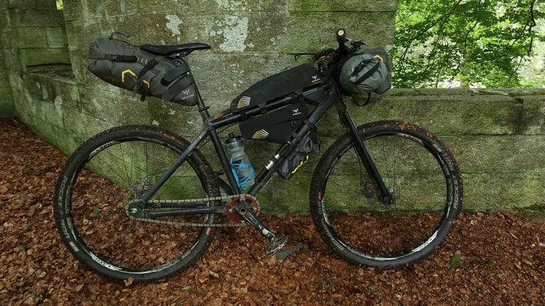 Markus Stitz's singlespeed with Apidura bags and a Downtube Pack on the down tube, leaning on a mossy stone wall.