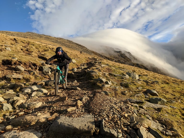 Beth Hodge rides a mountain bike down the rocky Rangers Path on Snowdon in winter sun, cloud over the ridge.