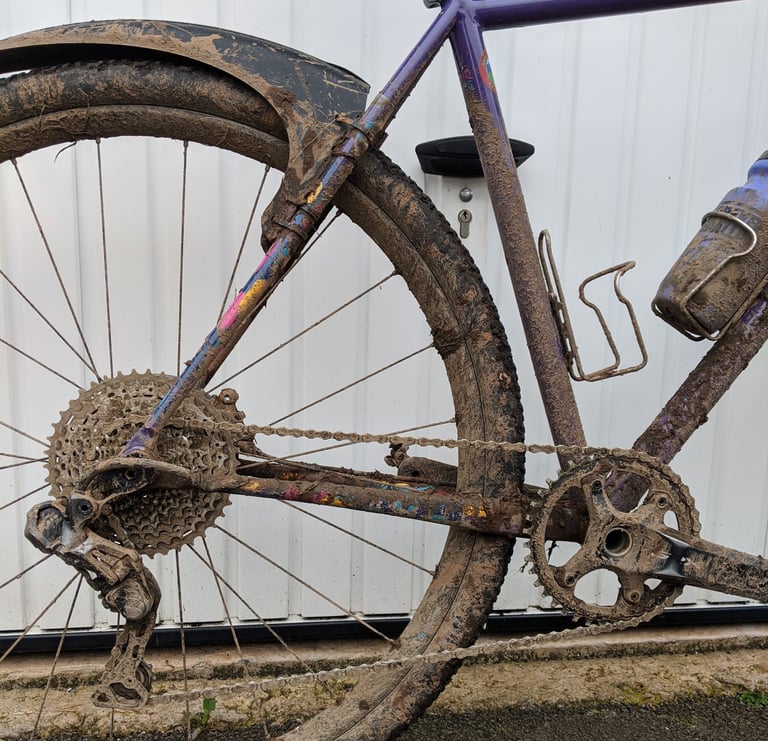 Gravel bike with a clip-on rear mudguard, wheel, chain and cassette caked in mud against a white garage door.
