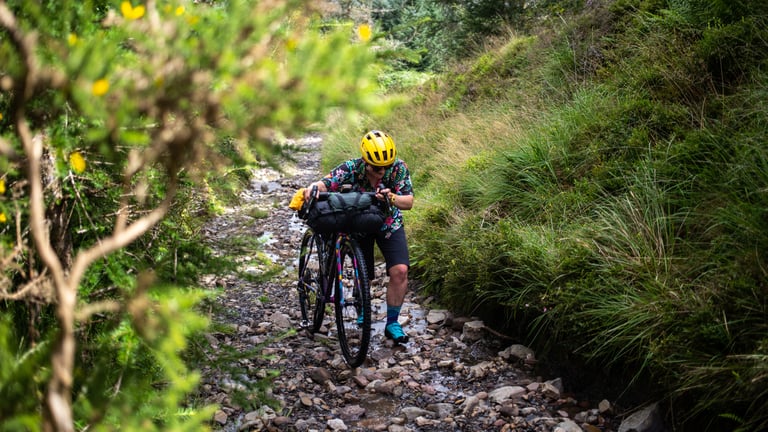 Rider pushes a loaded gravel bike up a rocky, water-washed track on Sarn Helen in South Wales.