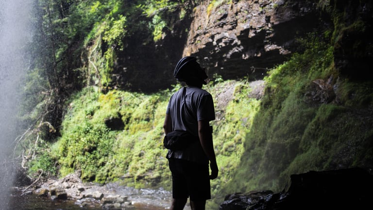 Silhouetted rider in a helmet looks up at the rock face from behind the spray of Henrhyd Falls, South Wales.