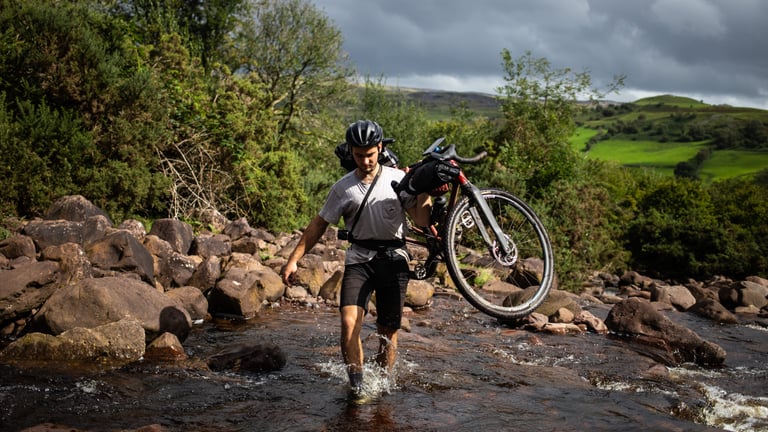 Chris Lord of Lord Cycles carries a steel bike on his shoulder across a rocky stream in the Welsh hills.