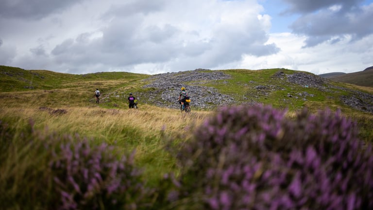 Three bikepackers cross open heather moorland under a cloudy sky on the Black Mountain, South Wales.