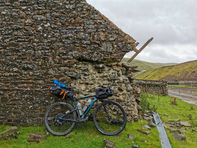 Molly Weaver's loaded bikepacking bike leaning against a ruined stone barn below grassy hills on the GB Divide.