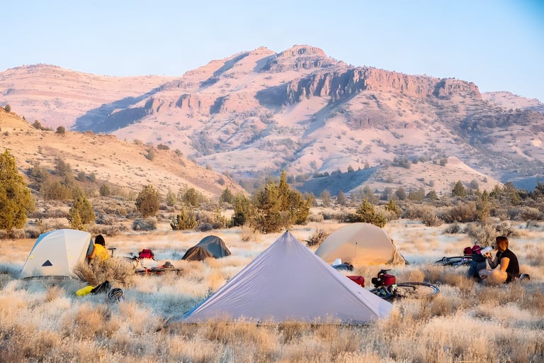 Bike campers' tents and loaded bikes in dry grass below rocky desert hills in low sun, ahead of the Swift Campout.