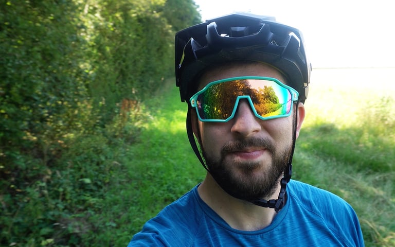 Smiling rider in a helmet wearing SunGod Velans glasses with mirrored Gold 8KO lenses beside a grassy path.