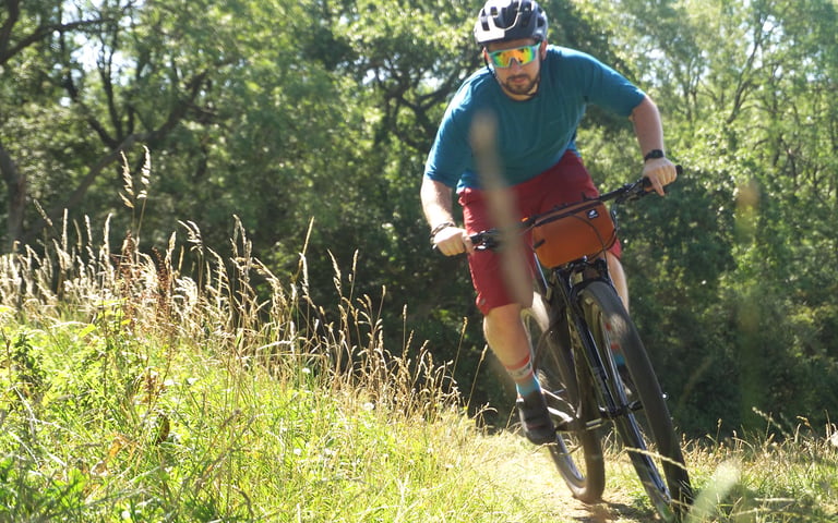 Rider wearing SunGod Velans glasses corners on a grassy off-road trail on a mountain bike with a handlebar bag.