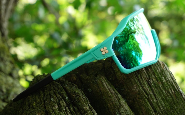 Minty-green SunGod Velans frame with mirrored lens and gold emblem on the arm, resting on a tree stump.