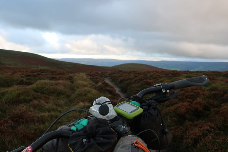 Handlebar bags and GPS unit on a bikepacking bike looking along a narrow trail over heather moorland at dusk.
