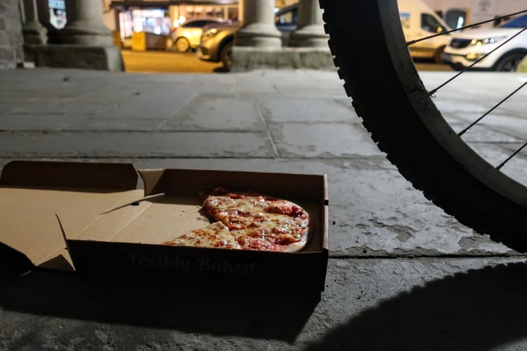 Half-eaten pizza in a box on a pavement at night beside a bike's tyre, a Bear Bones 200 refuel.