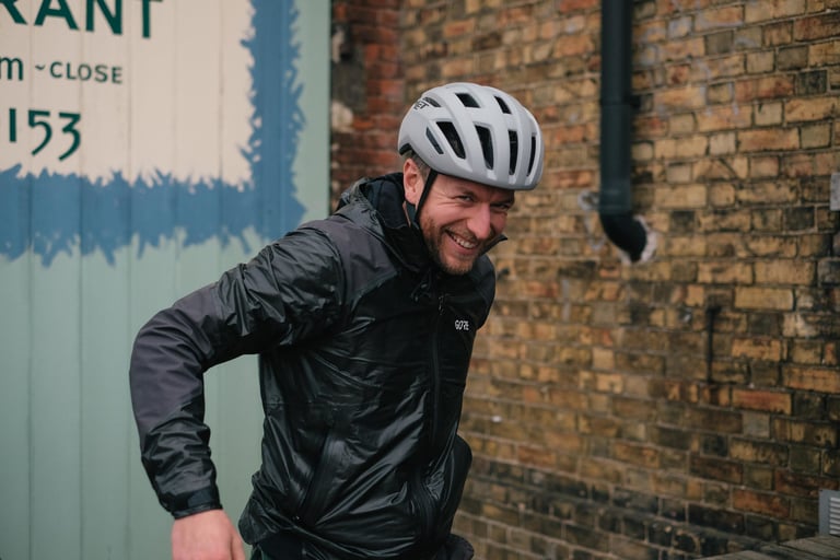 Will Meyer from Brother Cycles grins in a white helmet and rain jacket by the painted sign at The Goods Shed, Canterbury.