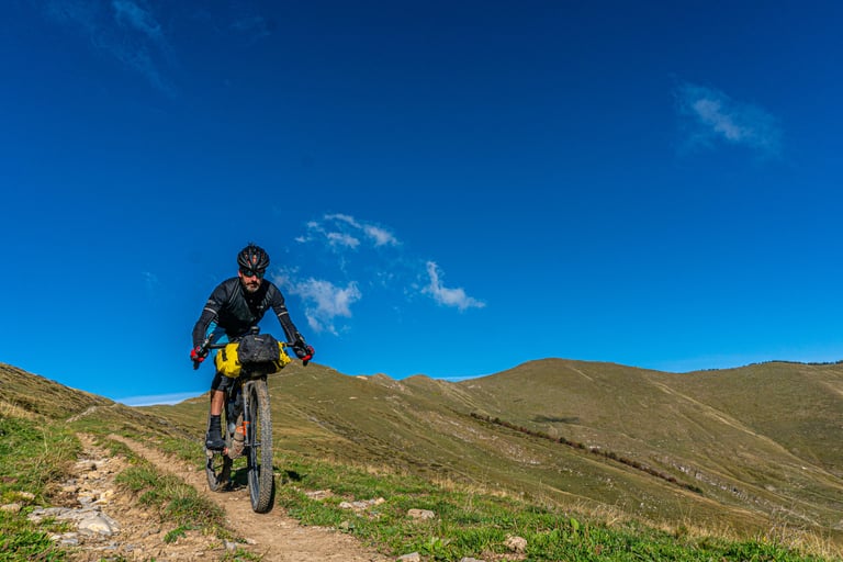 Scotty rides a bikepacking bike with Curve Walmer 55cm bars along a grassy ridge trail under a deep blue sky.