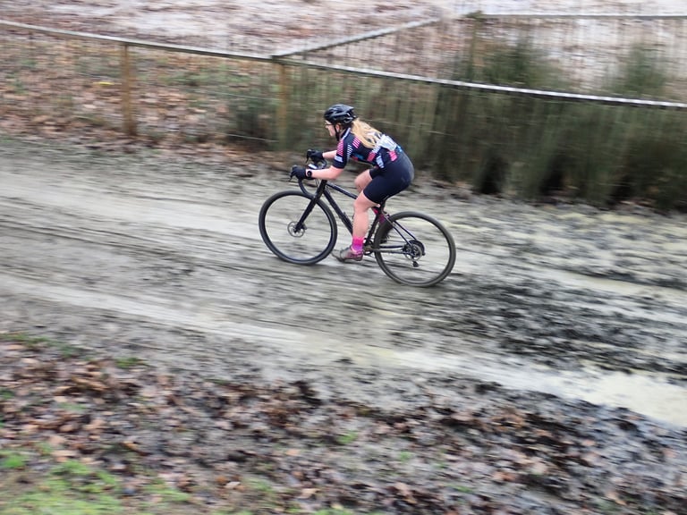 Rider on WTB Nano tyres speeds a gravel bike along a wide, wet and muddy track beside a wooden fence.