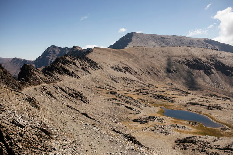 Gravel path traverses the high Sierra Nevada near Pico del Veleta, with a small lake below the summit.