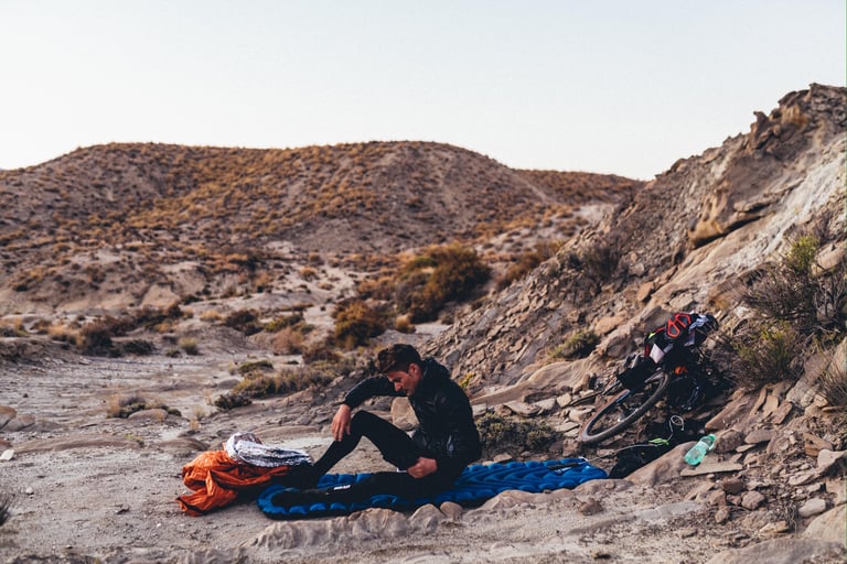 Badlands rider sits on a sleeping mat in a dry gully in the Tabernas desert, with a bike propped on the rocks.