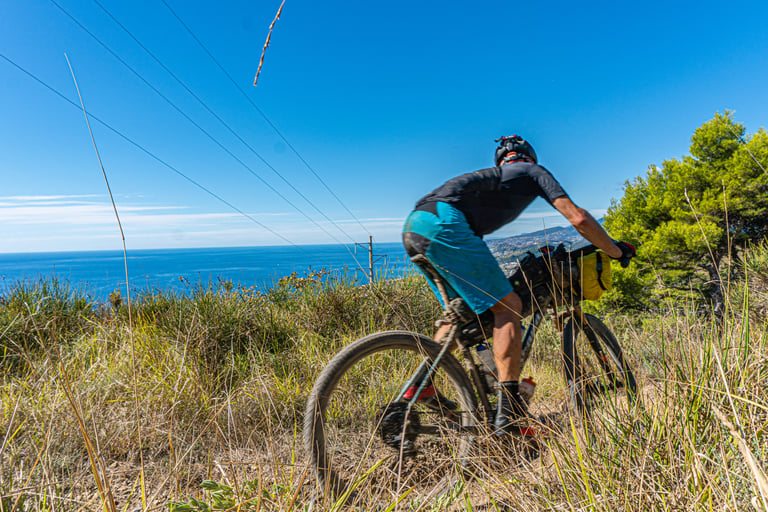Rider on a Walmer-barred bikepacking bike drops down a grassy trail on the Ligurian coast with the sea below.