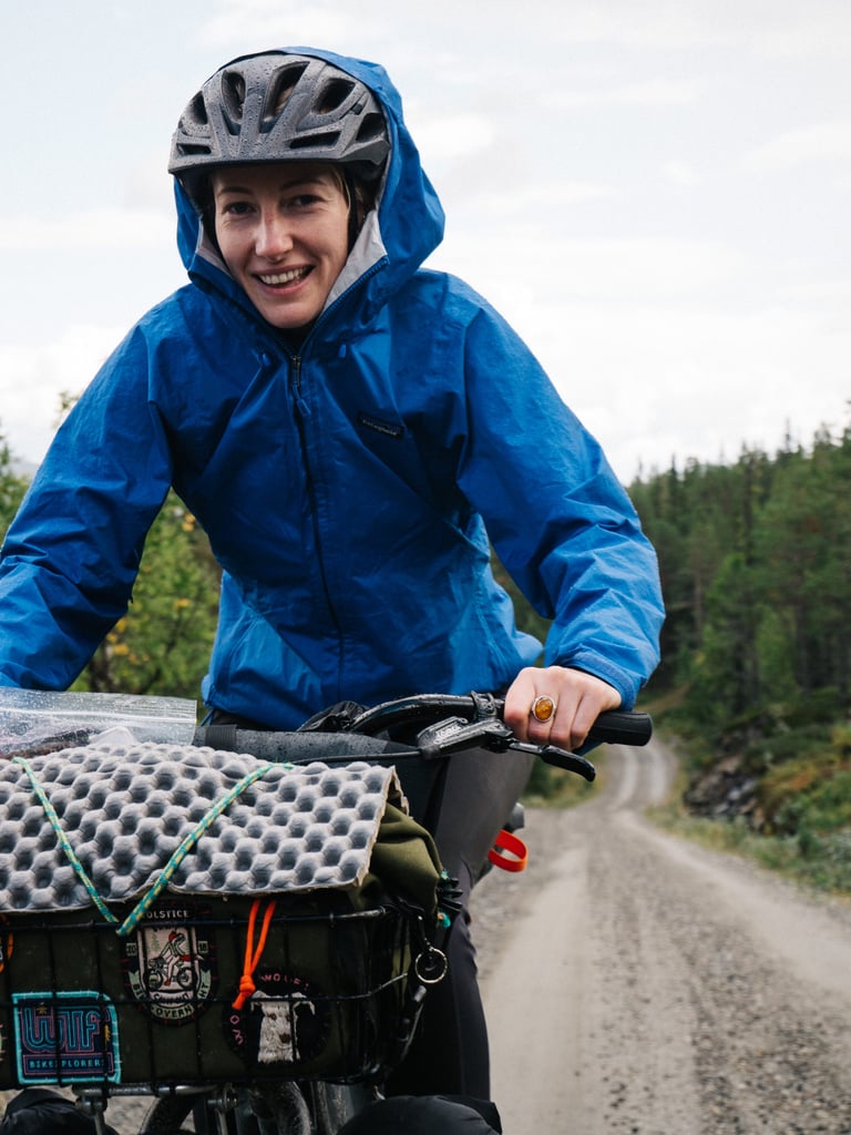 Smiling rider in a blue rain jacket on a gravel road, with a foam mat strapped to a front basket.