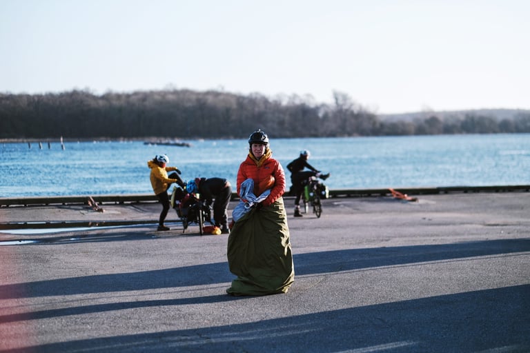 Resolution Race rider in an orange jacket stands in a bivvy bag beside the water while teammates pack their bikes.