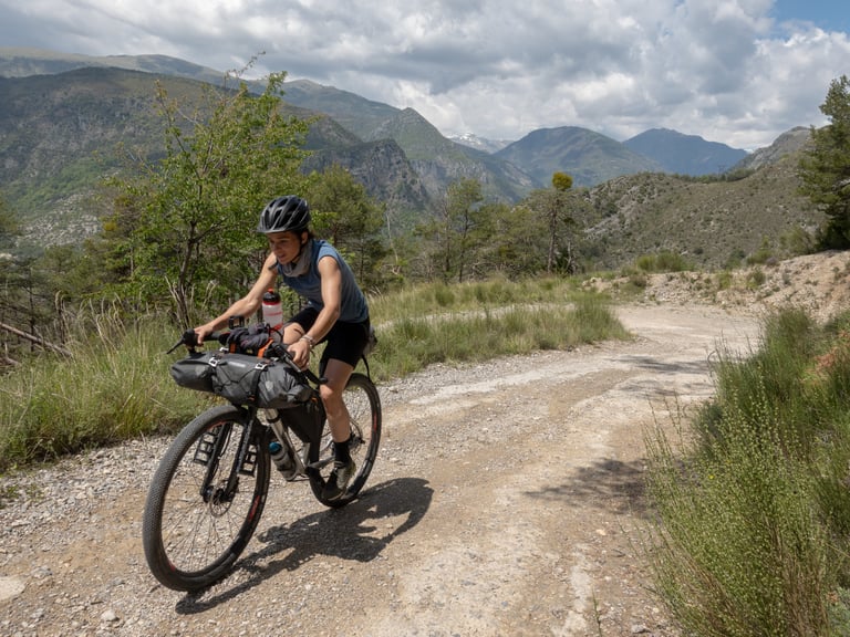 Rider on a Riverside touring bike with bar bag climbs a gravel track in Mercantour National Park, southern France.