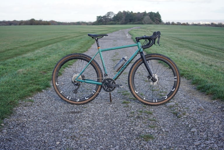 Stayer Groadinger UG test bike in teal with Stayer carbon wheels, drive side, on a gravel track across open grassland.