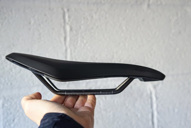Side view of the Fizik Terra Argo X3 gravel saddle showing its curved profile, held in a hand against a white wall.