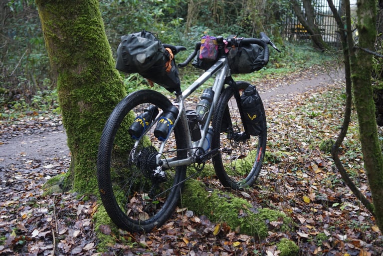 Loaded Riverside 920 with bar bag, seat pack and fork packs parked against a mossy tree beside a woodland track.