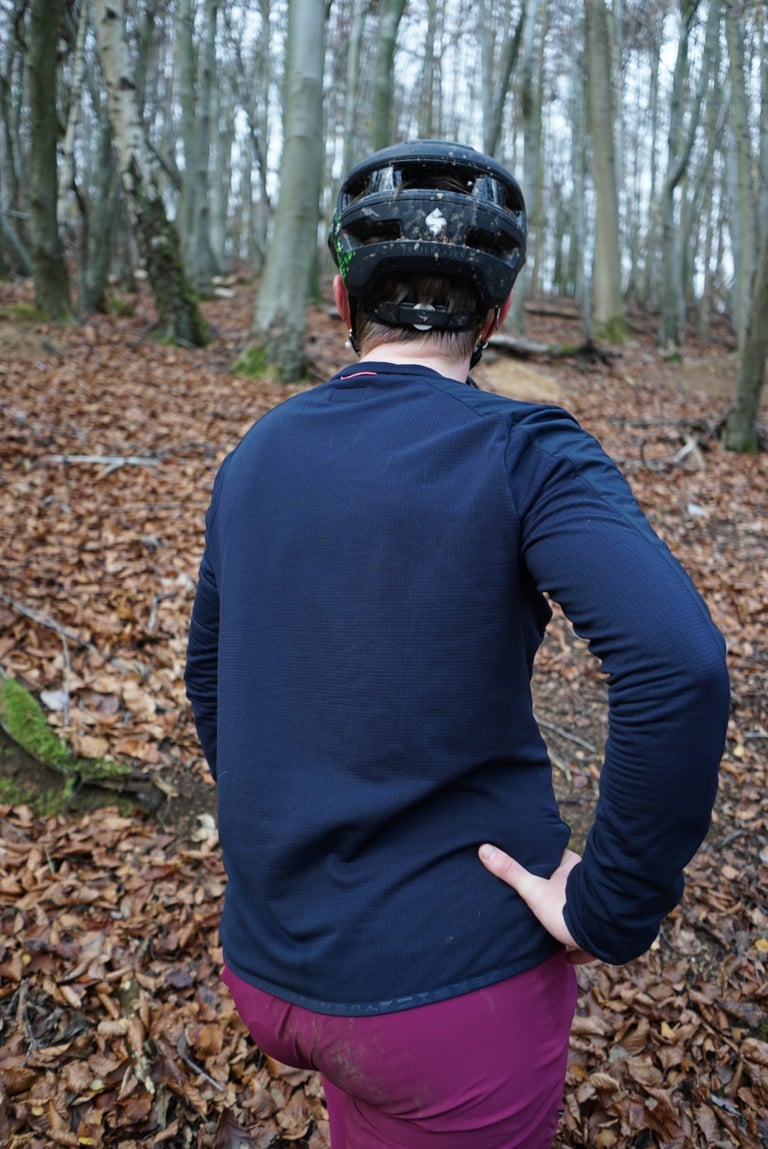 Back view of the loose-fitting Rapha Explore Long Sleeve Pullover in navy, worn with a muddy helmet in woodland.