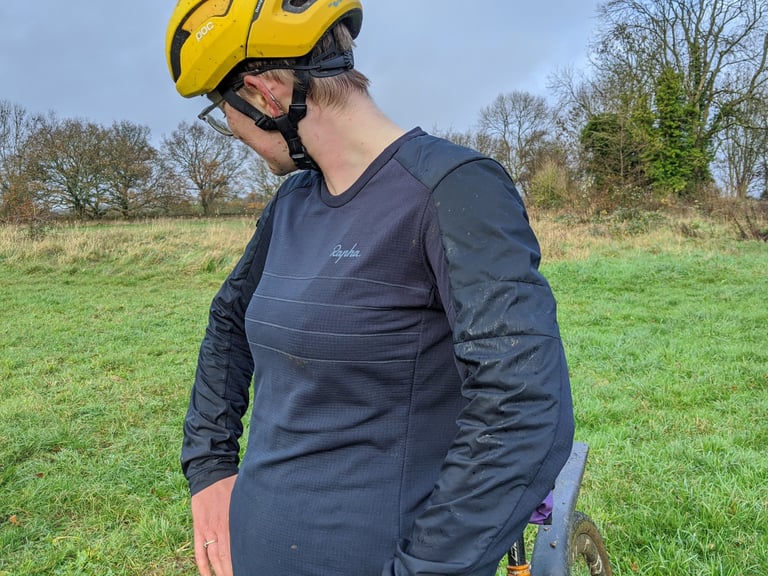 Navy Rapha Explore Long Sleeve Pullover with chest stripes and logo, worn by a rider in a field beside a bike.