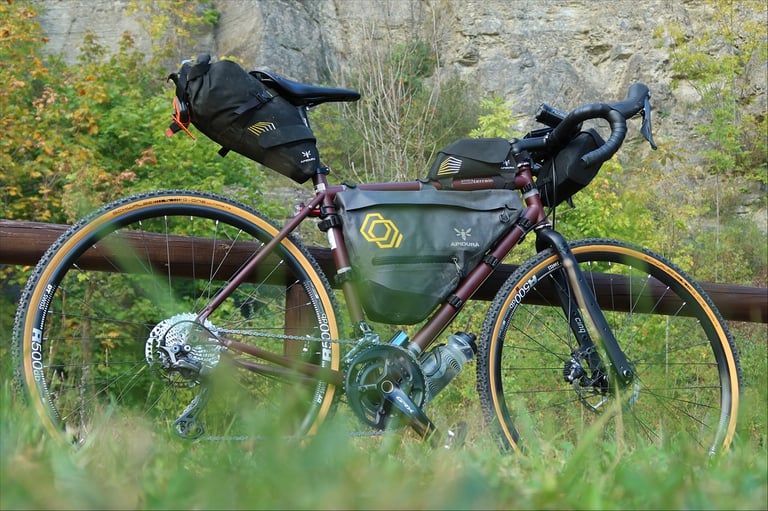 Markus Stitz's dark red Tout Terrain Vasco loaded with Apidura bags, leaning on a wooden fence below a rock face.