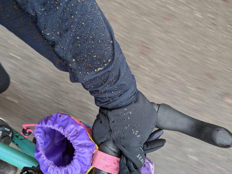 Water droplets bead on the windproof sleeve of the Rapha Explore pullover above a gloved hand on the handlebar.