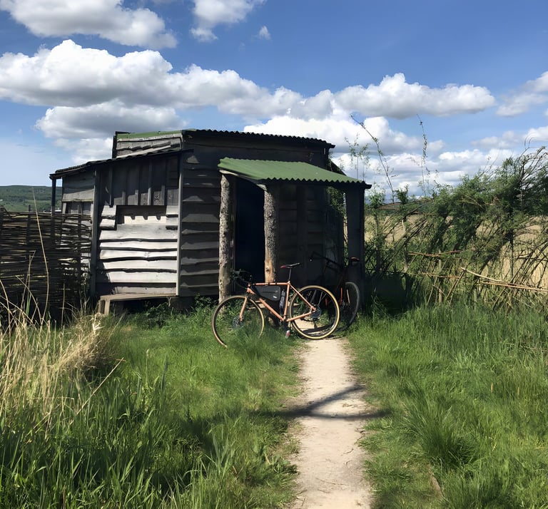 Gravel bike parked outside the weathered wooden Park End Moss bird hide on the Sizergh Estate, under a blue sky.