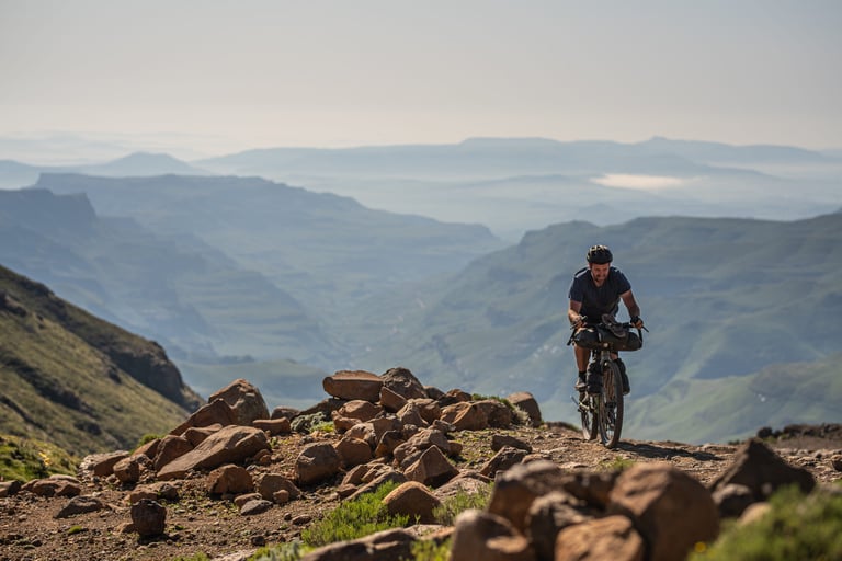 Rider on a loaded Riverside touring bike climbs a rocky track above misty valleys in Lesotho.