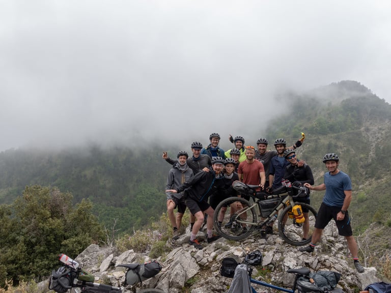 Riverside team and ambassadors in cycling kit pose with a loaded bike on a rocky ridge above misty hills.