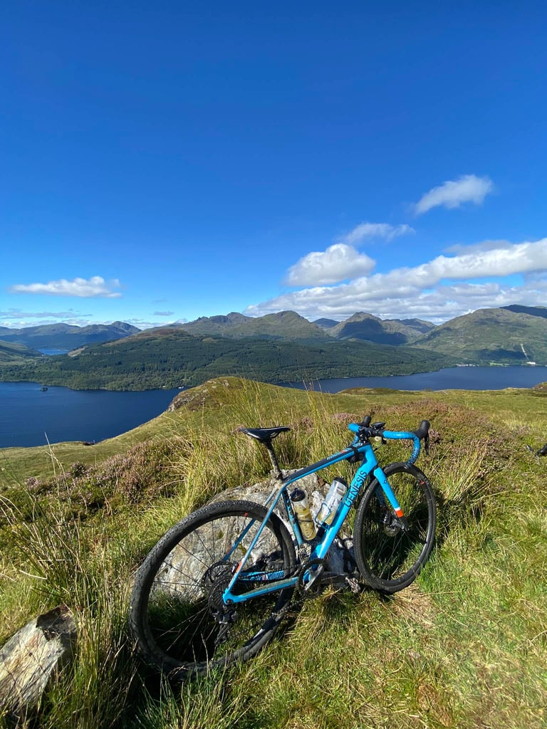 Blue Genesis gravel bike leans on a rock above a loch and mountains in the Gravelfoyle area of the Trossachs.