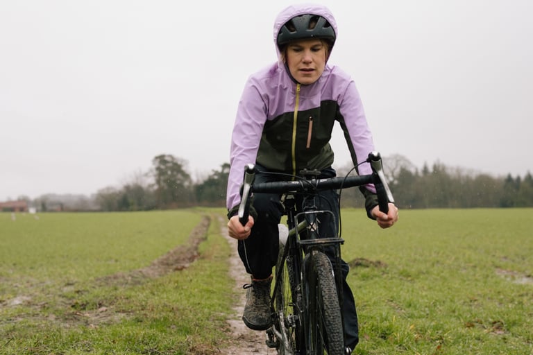 Winter test ride along a muddy field edge in the IRIS Escape Jacket, hood up over the helmet in drizzle.