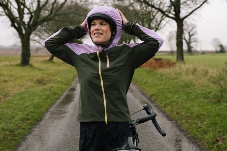 Rider pulls the lilac hood of the IRIS Escape Jacket up over a cycling helmet on a wet country lane.