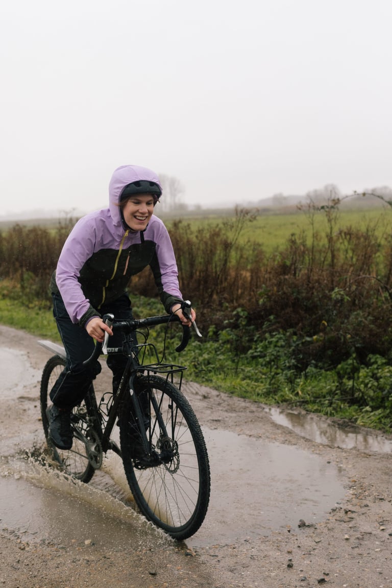Rider in the IRIS Escape Jacket with the hood up splashes through a muddy puddle on a gravel track.