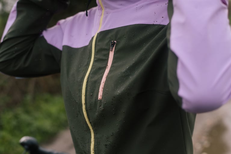 Close-up of the IRIS Escape Jacket's green front with water droplets, a yellow zip and pink chest pocket.