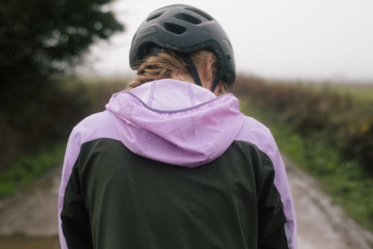 Back view of the IRIS Escape Jacket with its lilac hood resting below a helmet, flecked with rain.