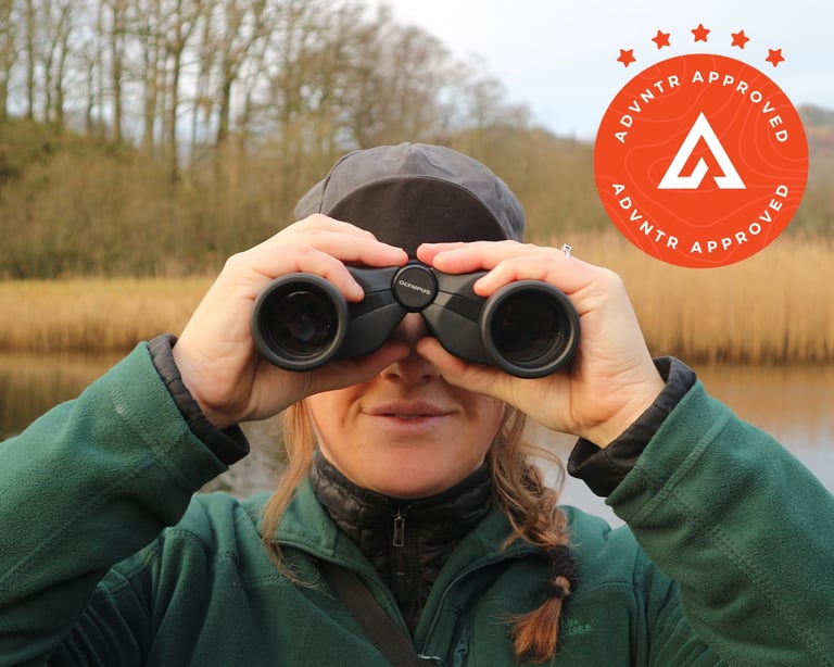 Person in a Rough Stuff Fellowship winter cap and green fleece looks through Olympus binoculars beside a reedy lake.