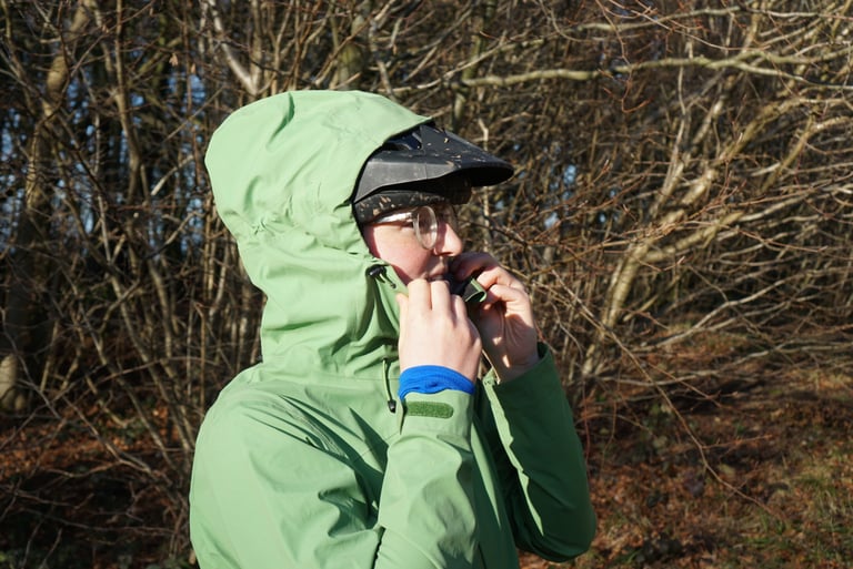 Rider pulls the Alpkit Balance hood up over a peaked helmet and adjusts the chin toggles, bare trees behind.