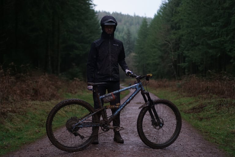 Mud-spattered rider in the Endura MT500 jacket, hood up, stands with a muddy blue mountain bike on a wet forest track.